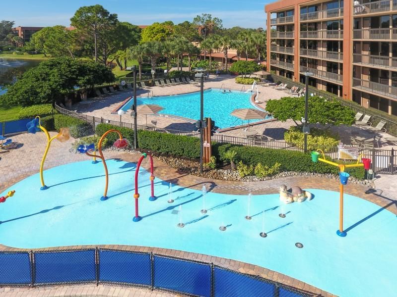 A large splash pad area with colorful fountain pipes, with a pool and hotel building just behind it. 