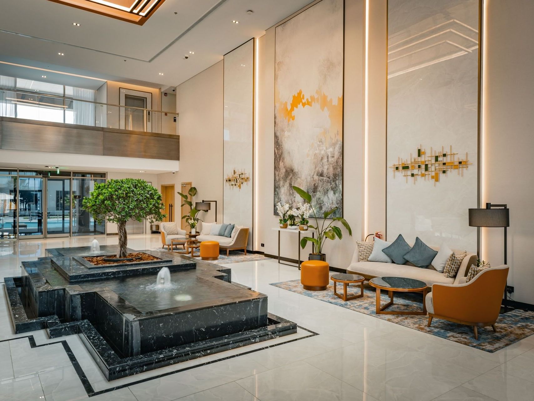 Modern lobby with a fountain, seating area, artwork, and plants, featuring a glass wall and balcony.