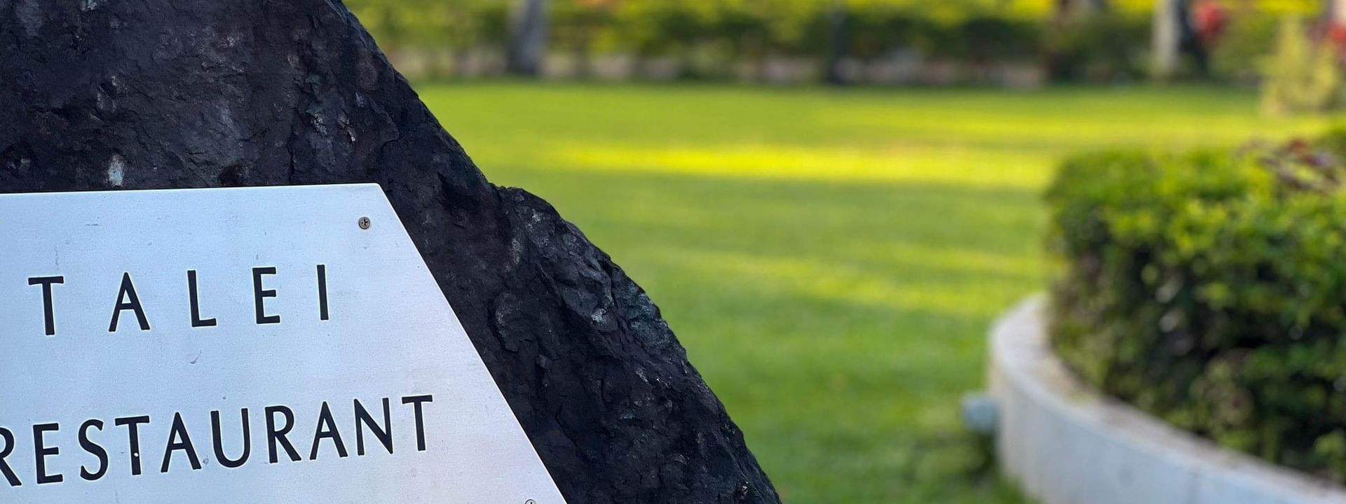 White Talei Restaurant sign by a dark volcanic rock surrounding a lush green lawn at The Naviti Resort in Korolevu