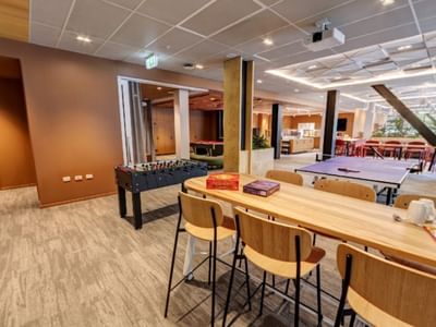 Modern communal lounge with foosball table, wooden seating, board games, and open dining area in Tupuārangi.