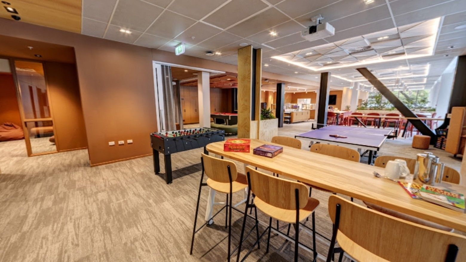 Modern communal lounge with foosball table, wooden seating, board games, and open dining area in Tupuārangi.