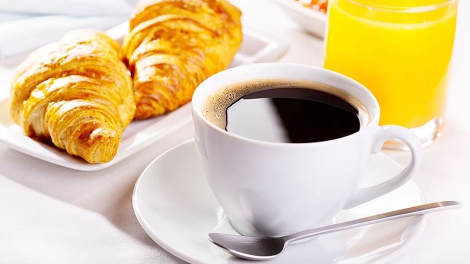 Freshly brewed cup of coffee next to two golden croissants & a glass of orange juice set on the table at The Diplomat Resort