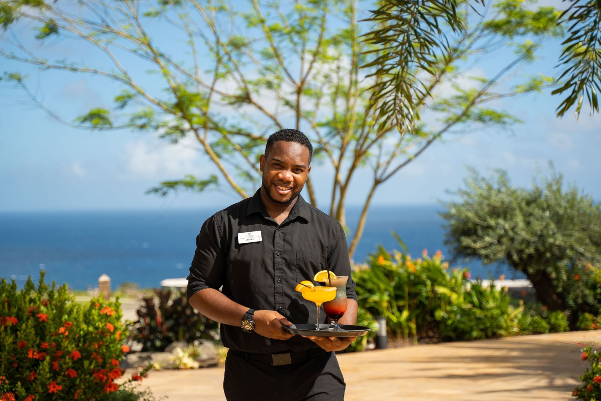 Waiter carrying a tray with colorful cocktails against a backdrop of greenery and a blue ocean at Golden Rock Resort
