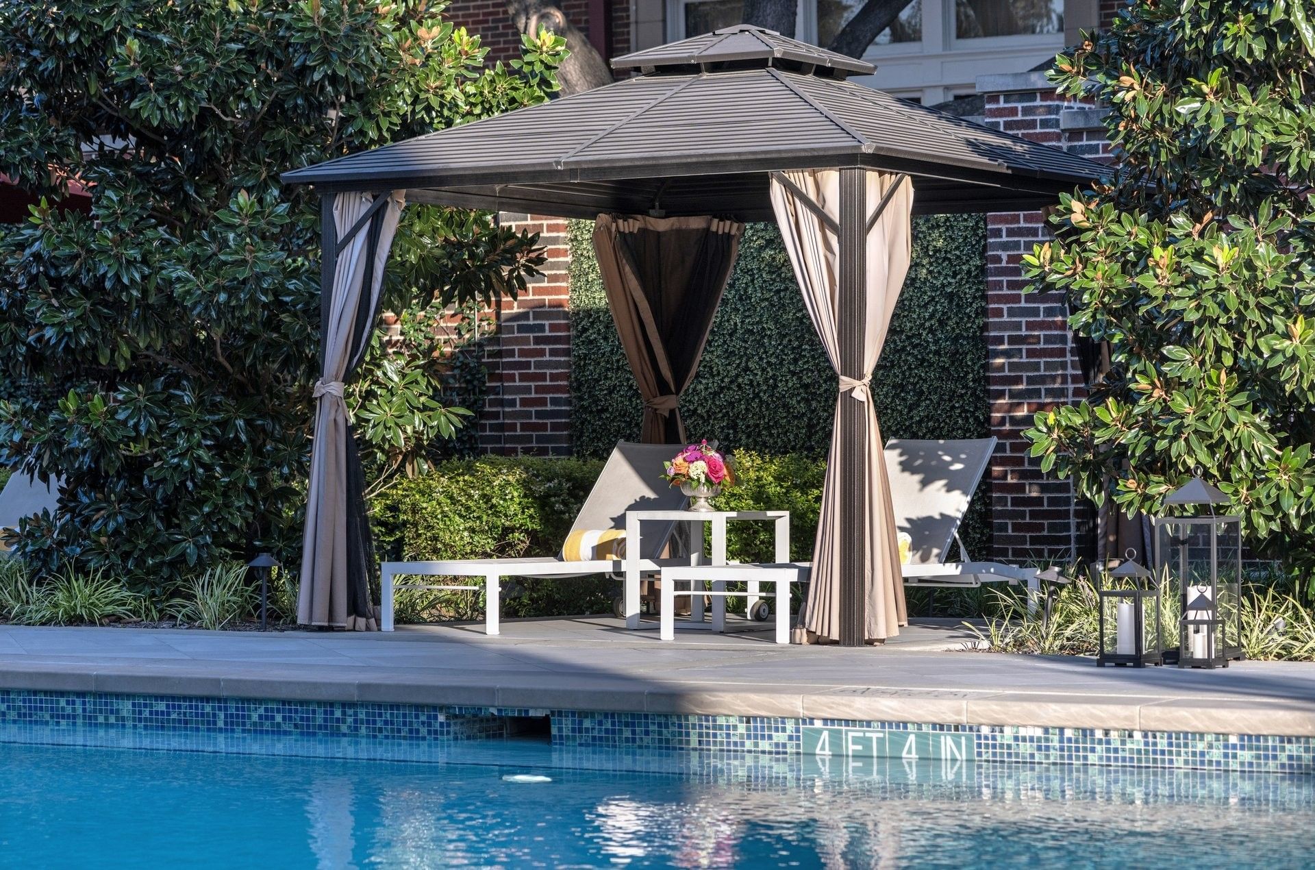 Lounge chairs by a table under a dark cabana surrounding the outdoor pool at Warwick Melrose Dallas