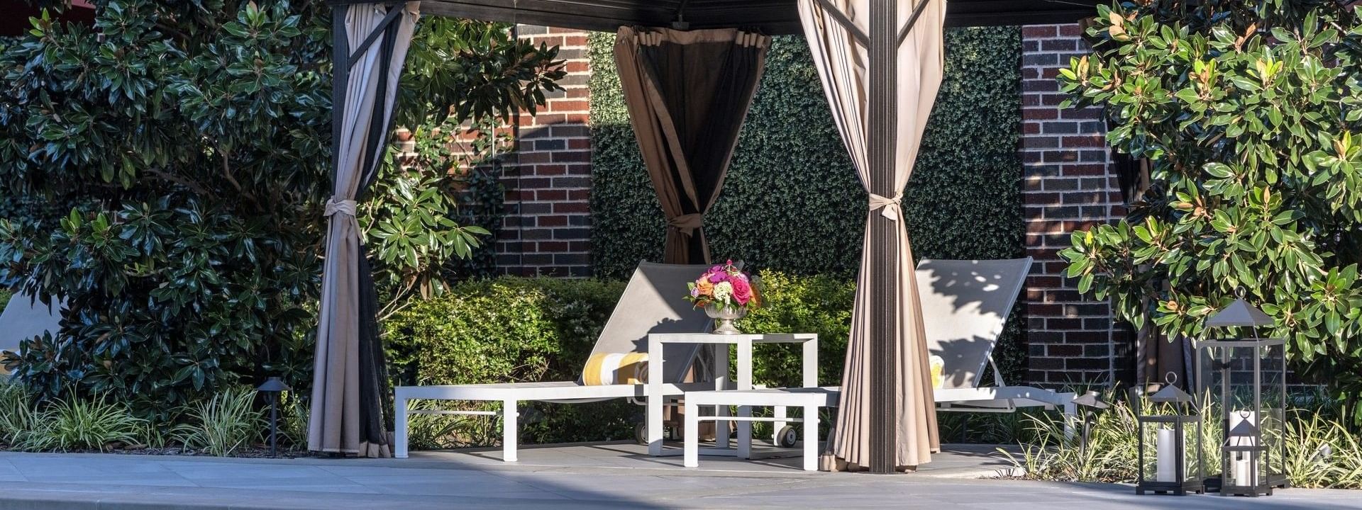 Lounge chairs by a table under a dark cabana surrounding the outdoor pool at Warwick Melrose Dallas