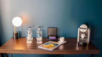 Wooden desk with lamp, coffee machine, bottles, cup, magazine, and electronic device against teal wall.
