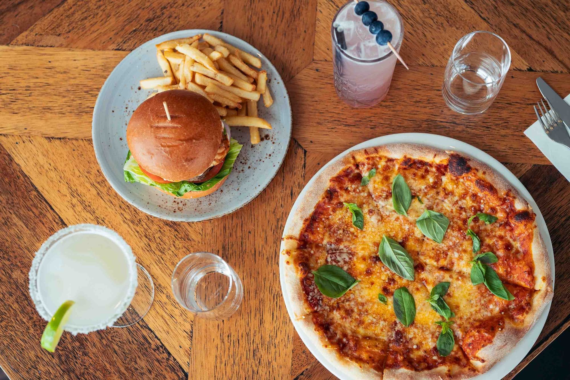 A burger, fries, pizza, and drinks on a wooden table with utensils and a napkin.