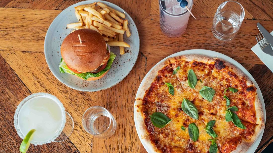 Wooden table with burger, fries, pizza, and drinks including a margarita with lime wedge.