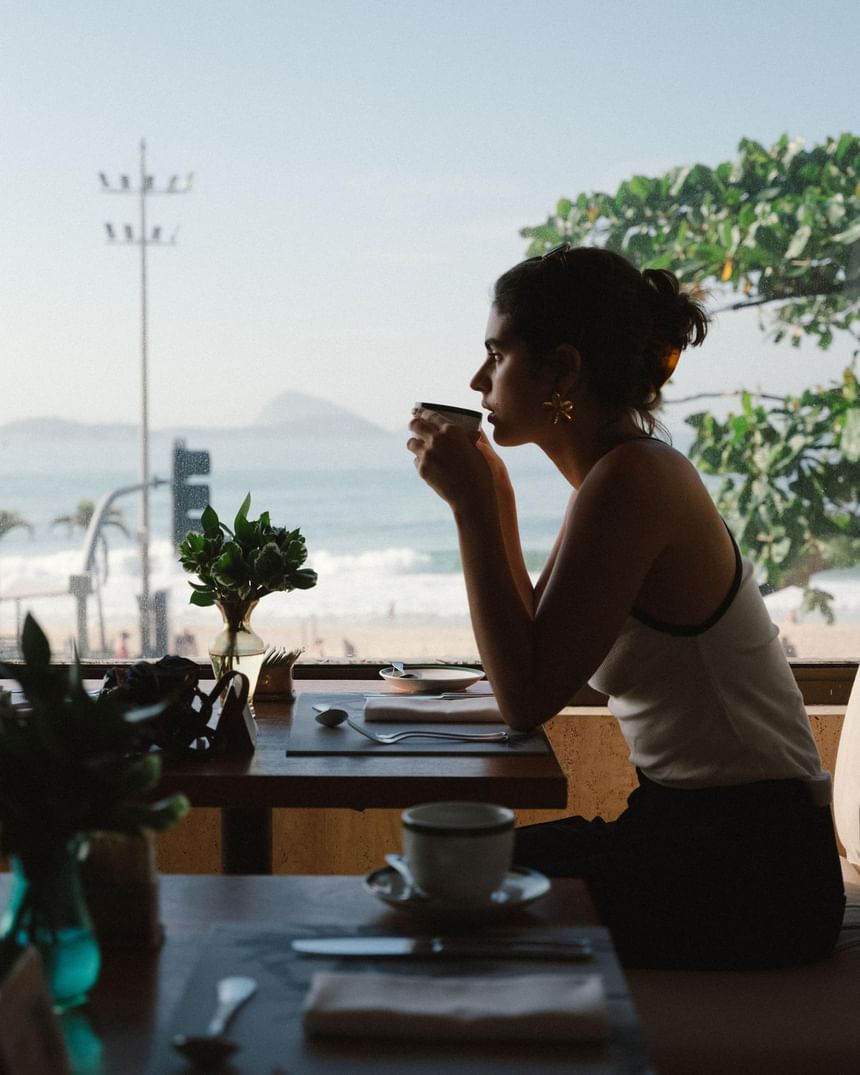 Uma mulher tomando café em uma mesinha de café da manhã do Janeiro Hotel de frente para o mar