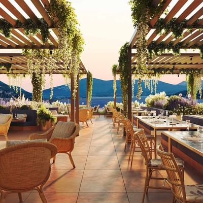 Ristorante esterno con pergolato, tavoli, sedie, bottiglia di champagne, viste su montagne e mare.