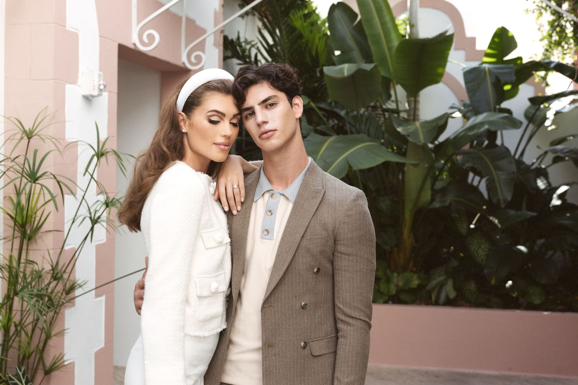 A couple stands in front of a pink and white building with tropical plants.