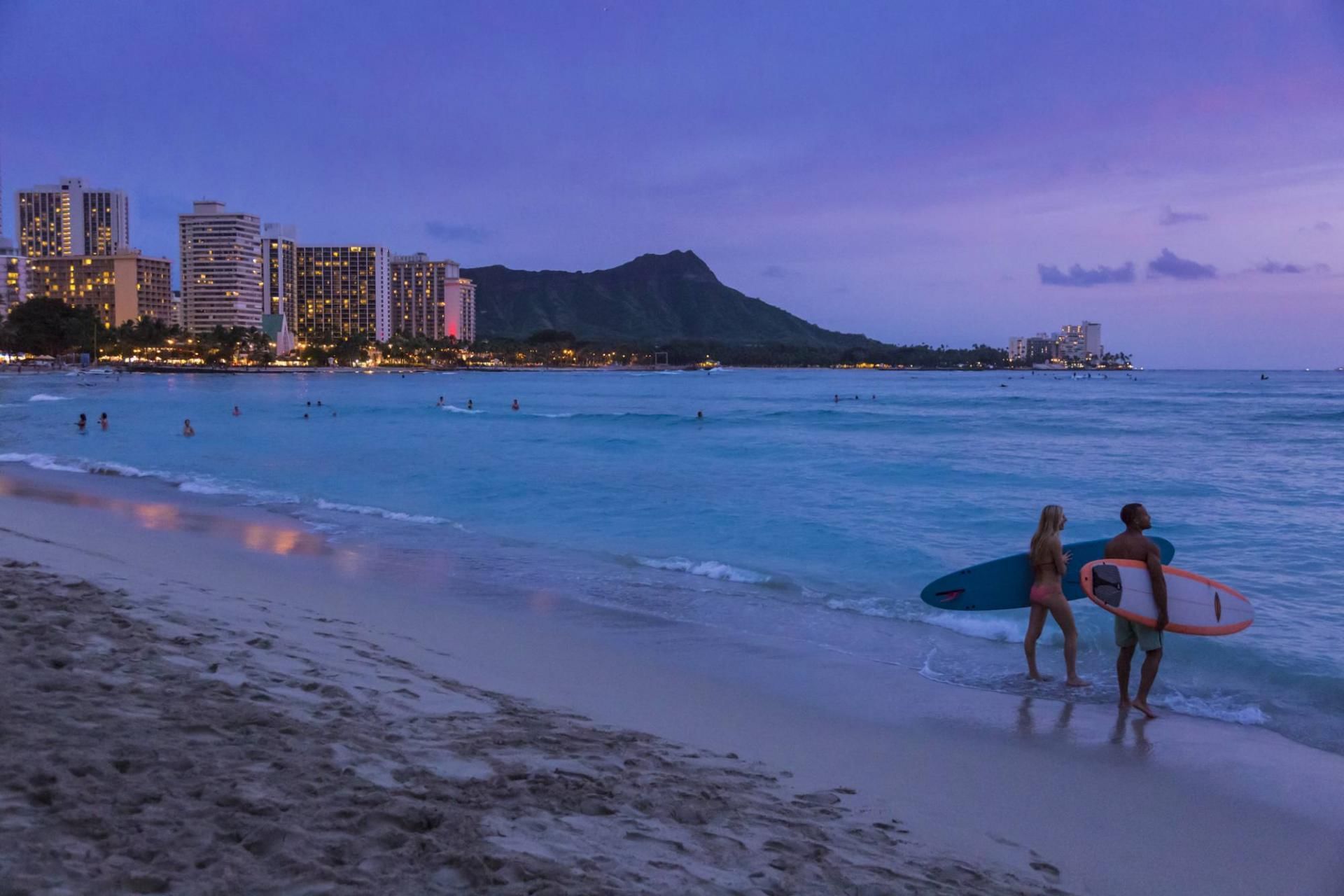 A beach scene at dusk with surfers and a city skyline in the background.