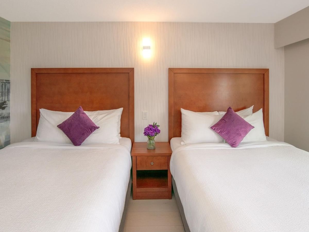 Two beds with white linens and purple pillows, a nightstand with flowers and a painting.