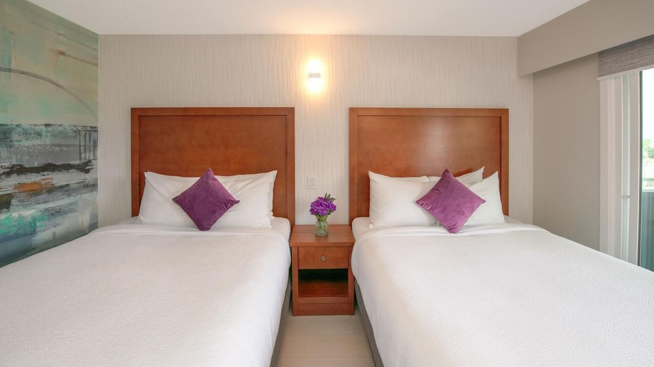 Two beds with white linens and purple pillows, a nightstand with flowers and a painting.