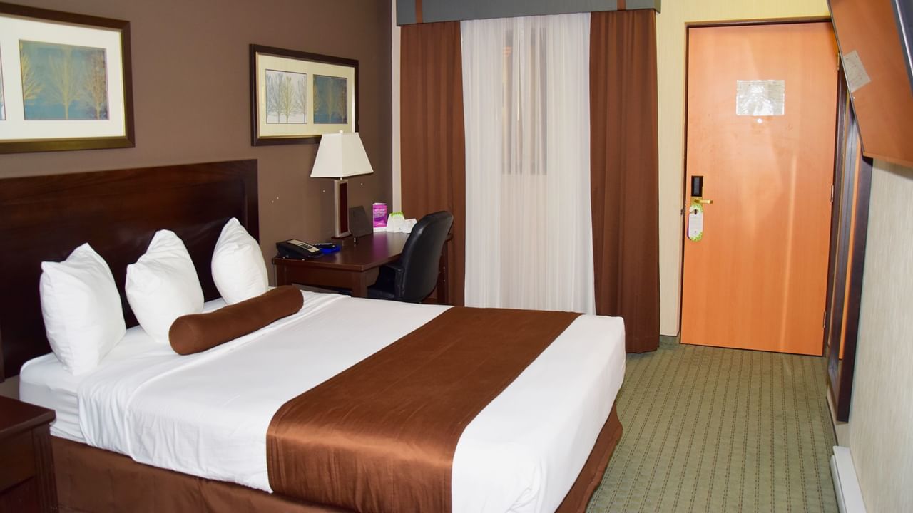 A hotel room with a bed, desk, lamp, and two framed pictures on the wall.