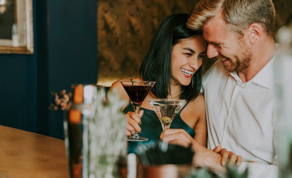 A man and woman at a bar lean into one another, laughing and holding martini glasses.