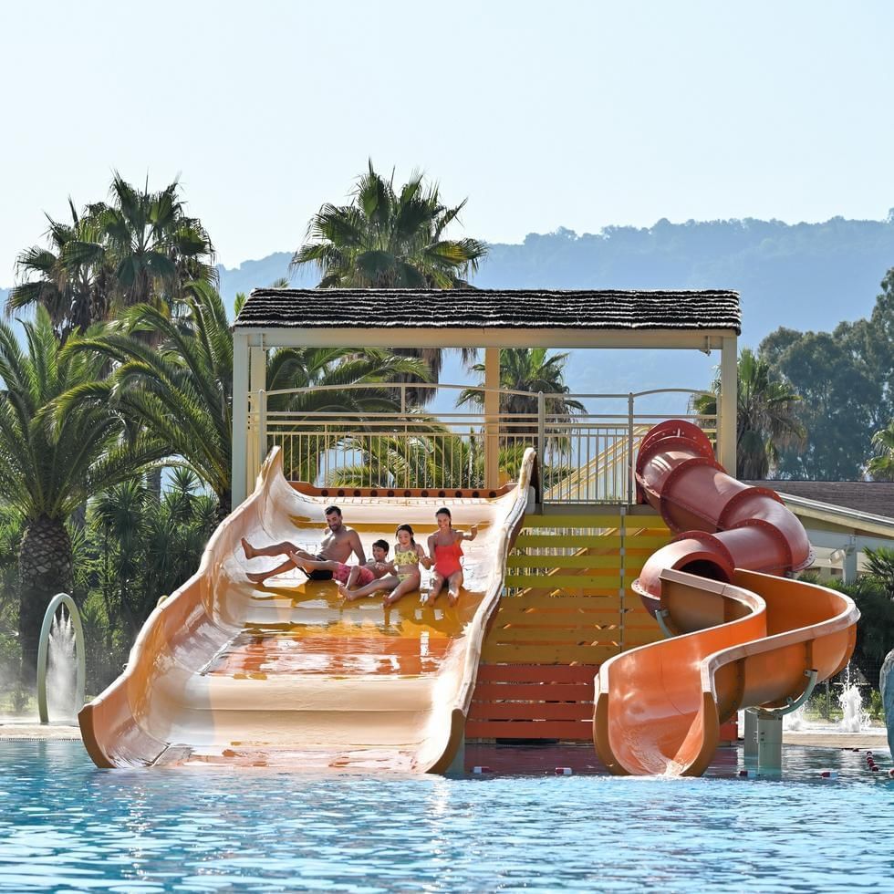 Familie auf Wasserrutschen im Schwimmbad, umgeben von Palmen und Hügeln im Hintergrund.