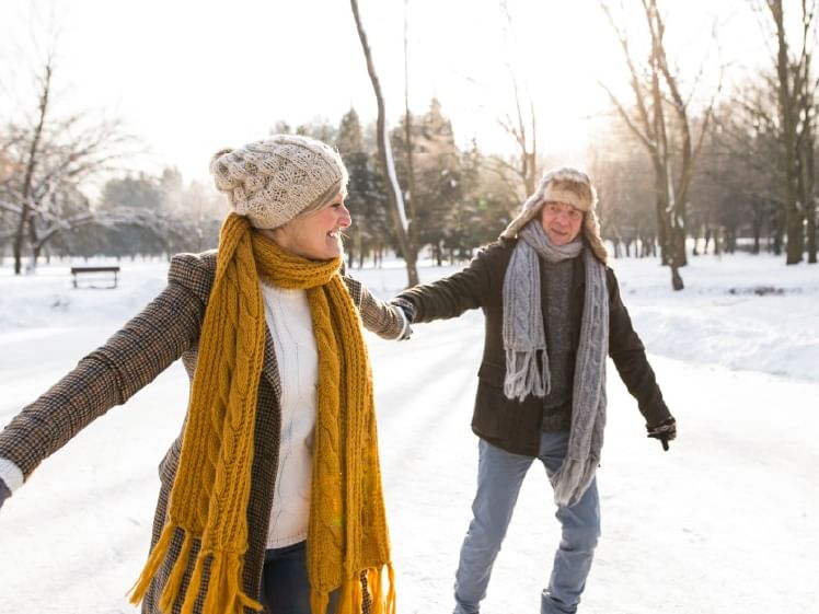 Two people ice skating together on a snow-covered path surrounded by trees and a park bench.