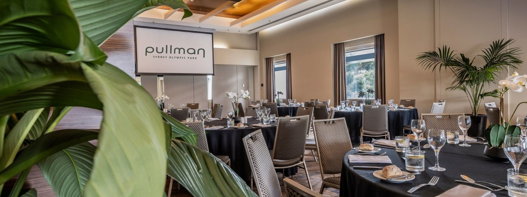 Elegant meeting room with black-clothed tables, wicker chairs, tropical plants, and natural light from large windows.