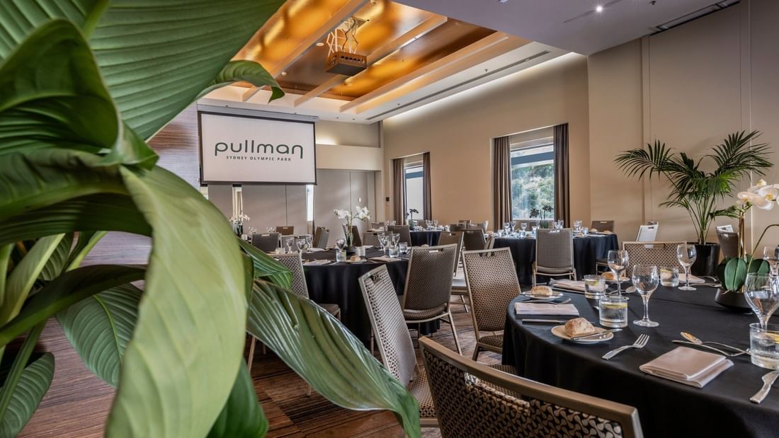 Elegant meeting room with black-clothed tables, wicker chairs, tropical plants, and natural light from large windows.