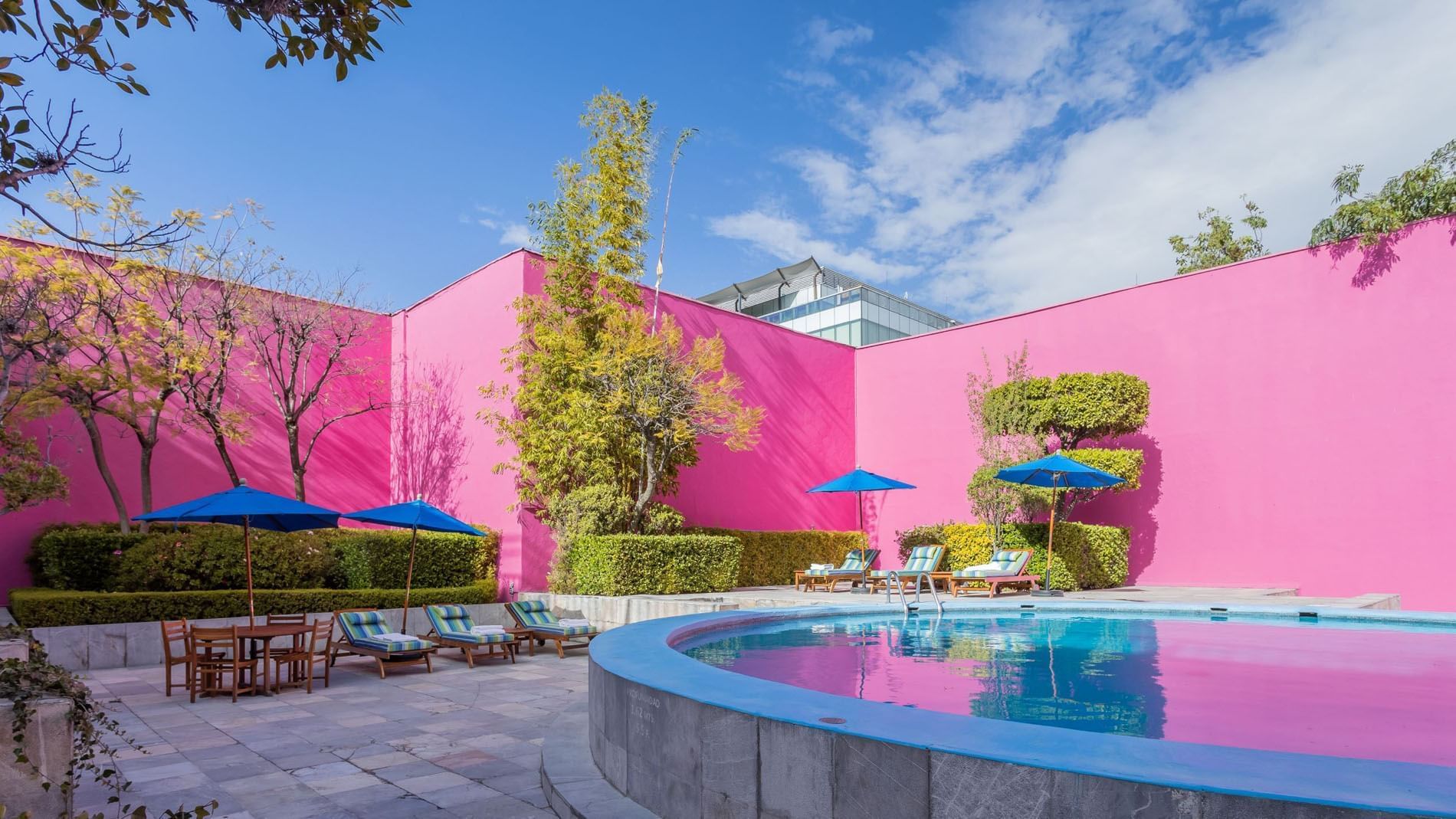 Piscina al aire libre con sillas, mesas y sombrillas azules junto a un muro rosa