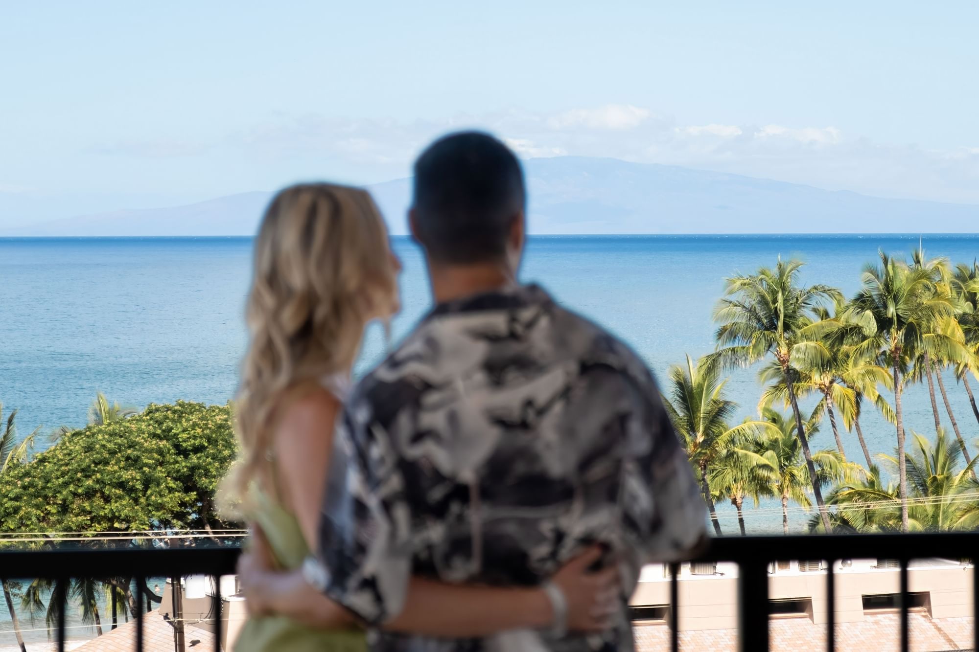 A couple is standing on a balcony overlooking the ocean and palm trees.