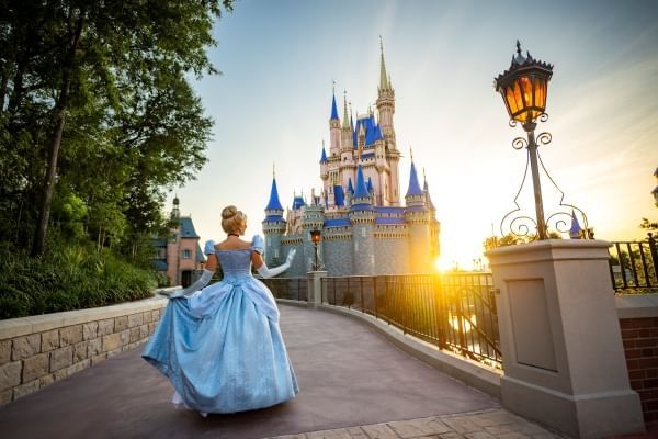 Cinderella in her classic blue dress stands on a path with her back to the camera facing the beautiful Cinderella Castle at sunrise. 