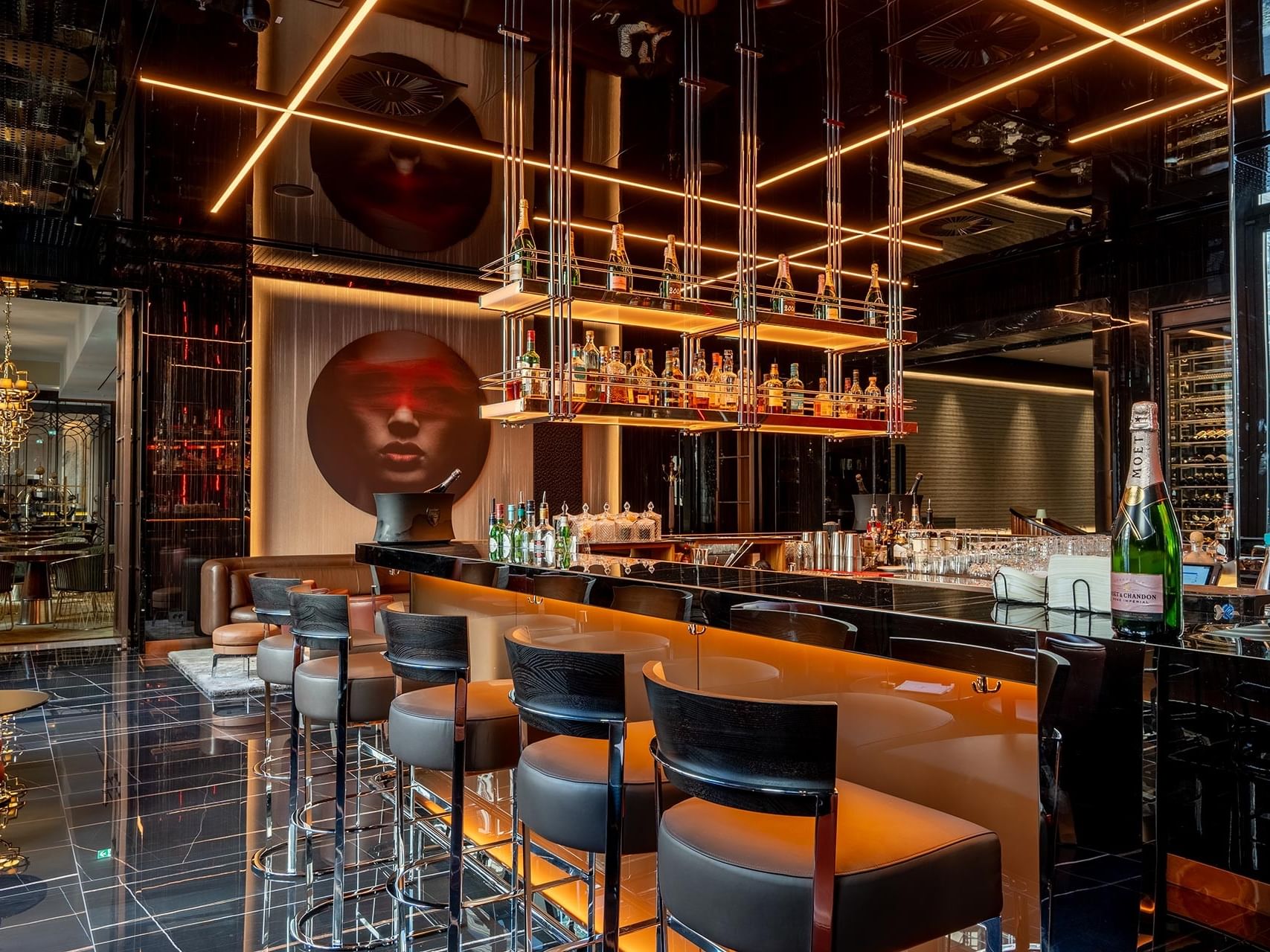 Modern hotel bar with orange curved counter, black leather stools, suspended bottle display, and artistic wall sculpture.