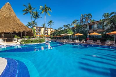 Resort pool with lounge chairs, umbrellas, and palm trees under a blue sky.