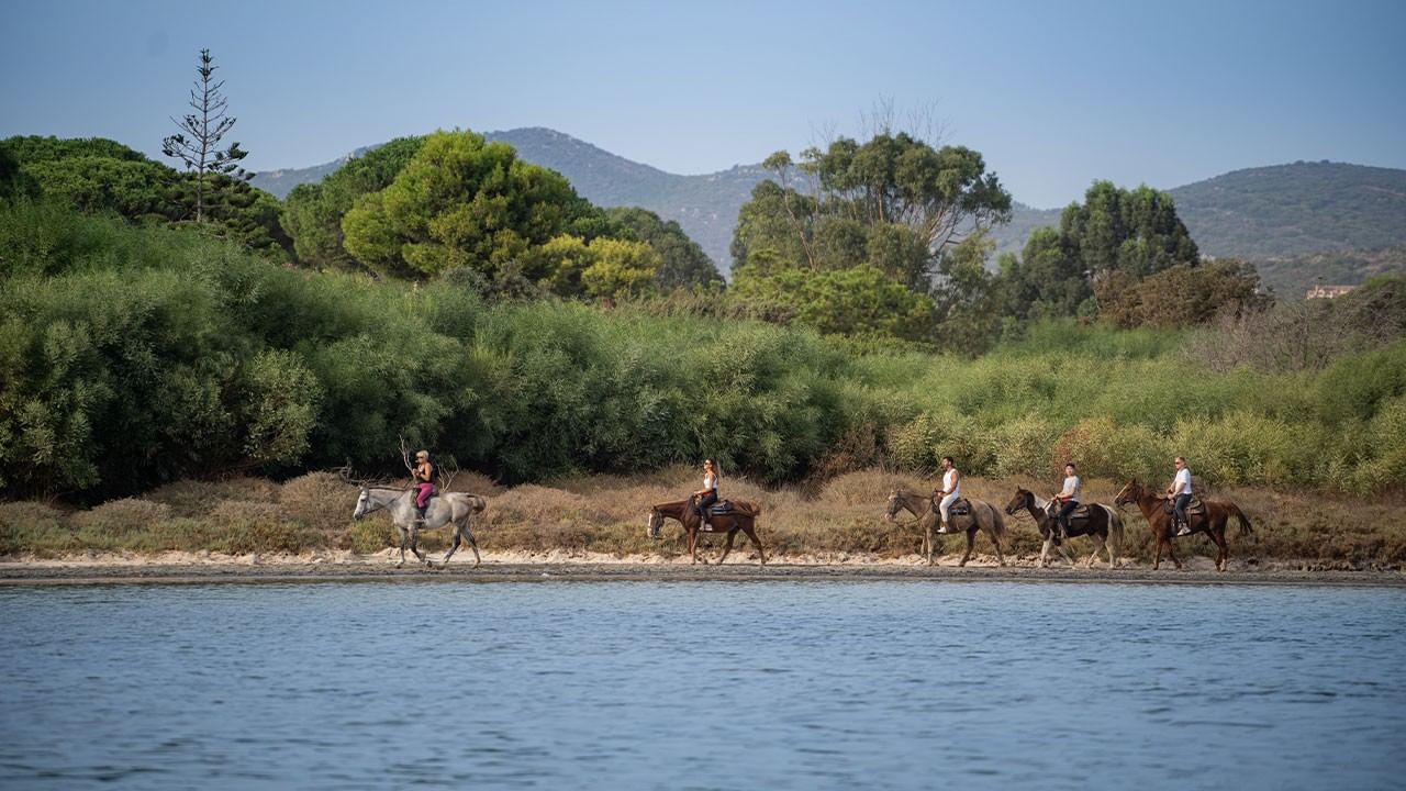 Gruppo di persone a cavallo lungo un lago con vegetazione e colline sullo sfondo.