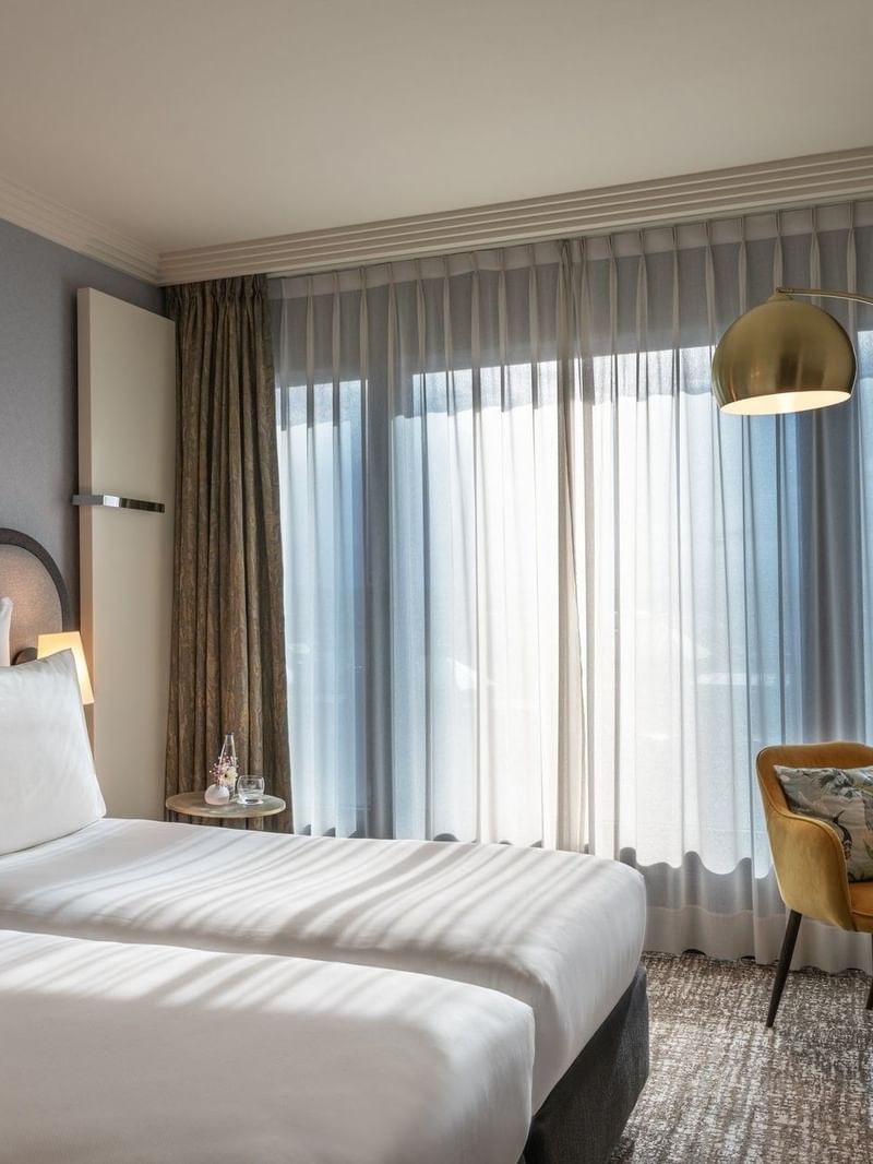 Twin beds by a yellow velvet armchair under a gold arched floor lamp near drapes in Classic Room at Warwick Geneva