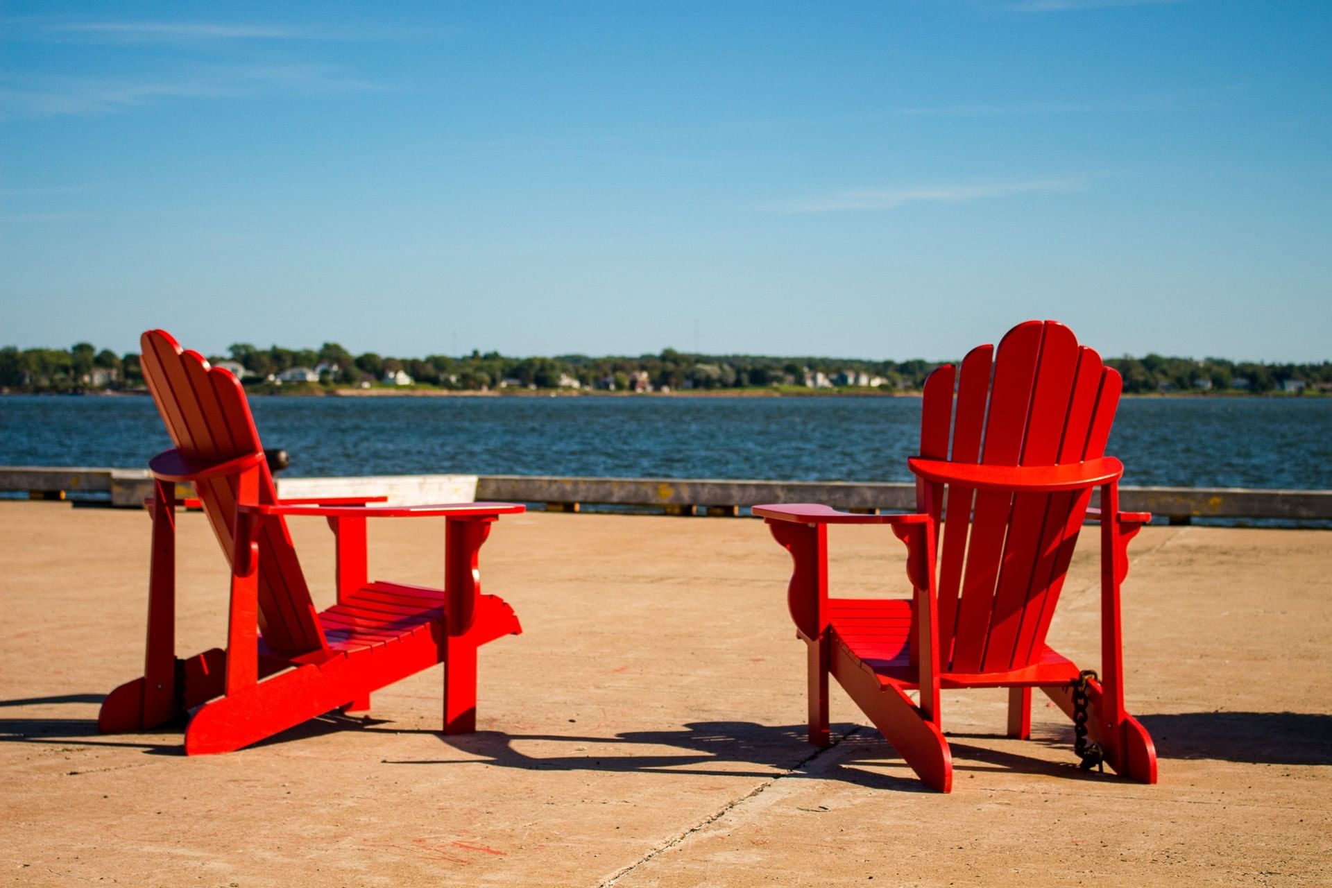 Two red Adirondack chairs on a concrete dock by a wide river under a clear blue sky at Rodd Charlottetown Hotel