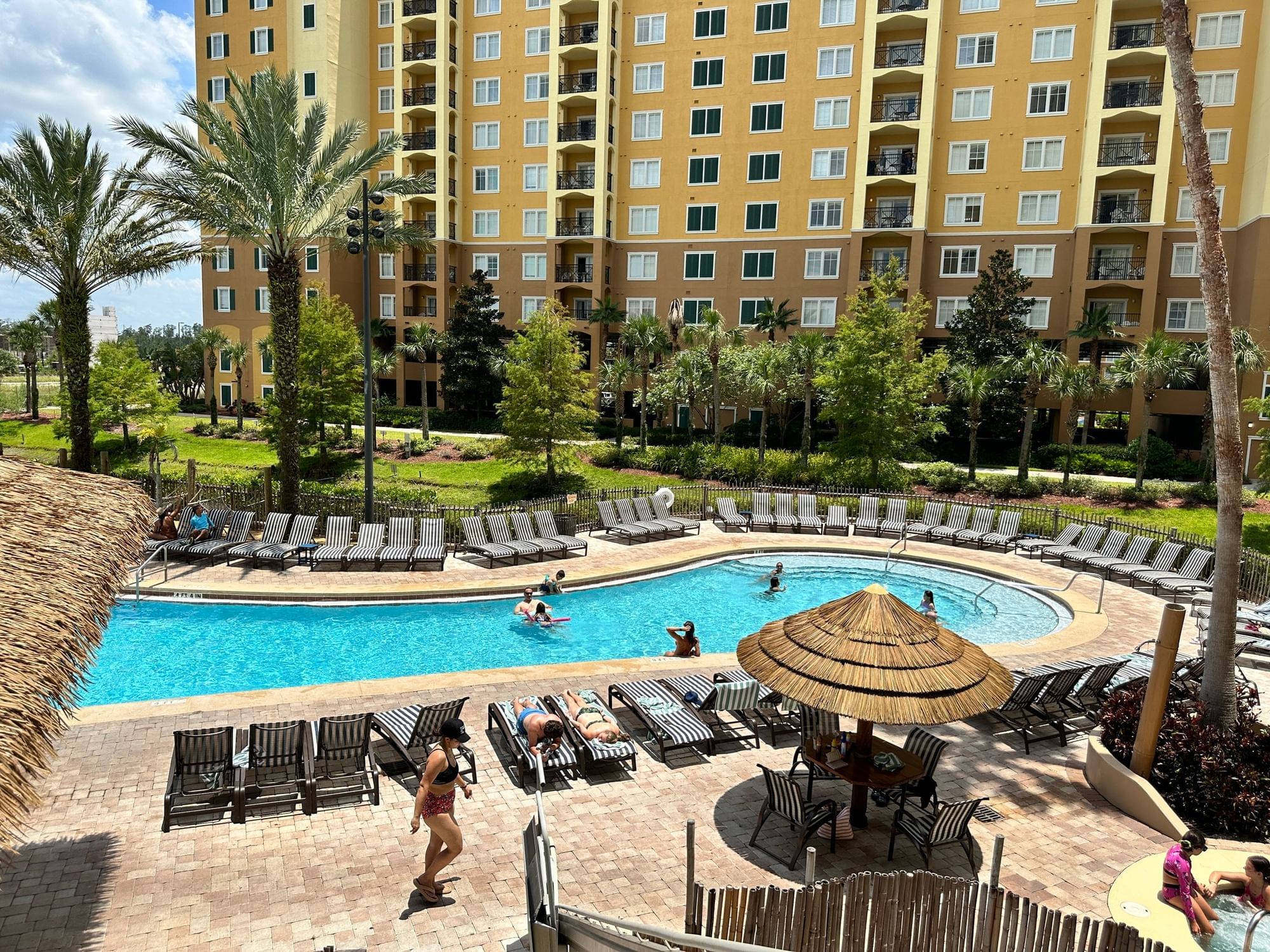 A large pool area with lounge chairs, palm trees, and a multi-story building in the background.