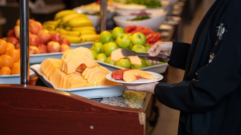 Person serve fresh sliced melon onto  white plate at breakfast buffet at Saja Warwick Madinah