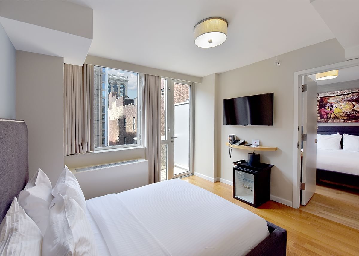 A hotel room with a large bed, window, TV, mini-fridge, and door to another room.