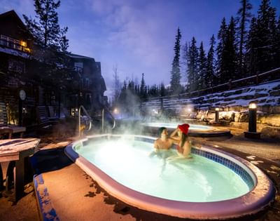 Couple relaxing in a steaming outdoor hot tub surrounded by snow-covered trees at Blackstone Mountain Lodge