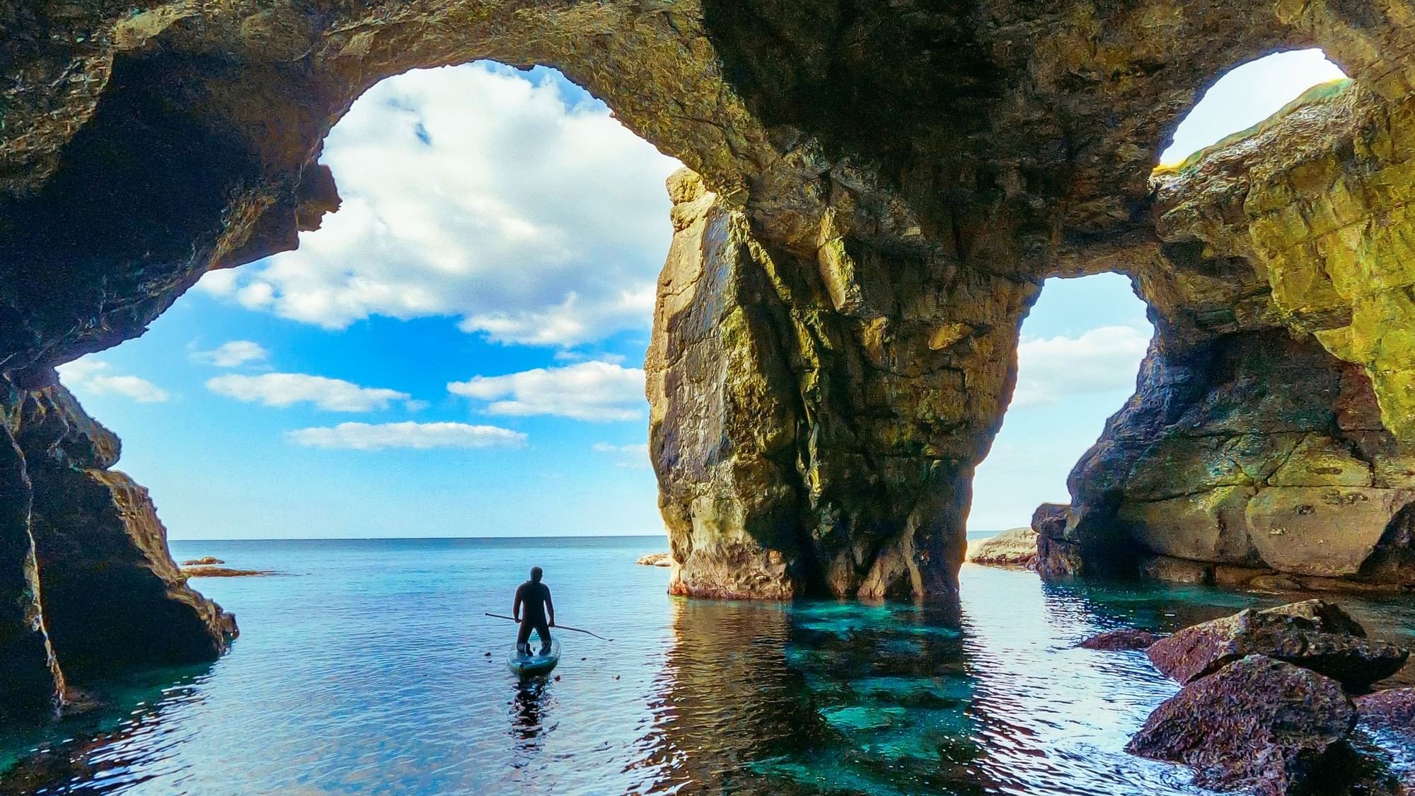 A person on a paddleboard explores a stunning sea cave with blue water and rock formations near Dunluce Lodge