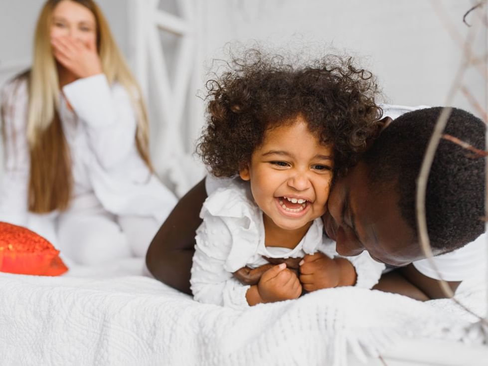 A young girl being adored by her dad on the hotel bed with mom giggling in the background.