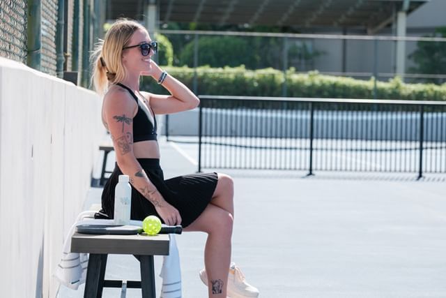 woman sitting on a bench at a tennis court with a tennis ball and water bottle
