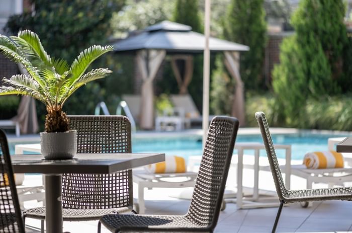Woven chairs by a dark table under a cabana near a sunlit swimming pool at Warwick Melrose Dallas