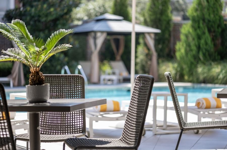 Woven chairs by a dark table under a cabana near a sunlit swimming pool at Warwick Melrose Dallas