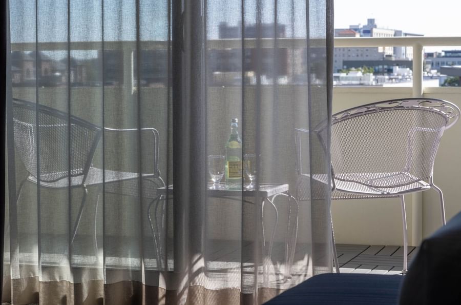 Balcony chairs by a table with water under a sunny sky through sheer curtains in Accessible Deluxe Rooms at Warwick Denver