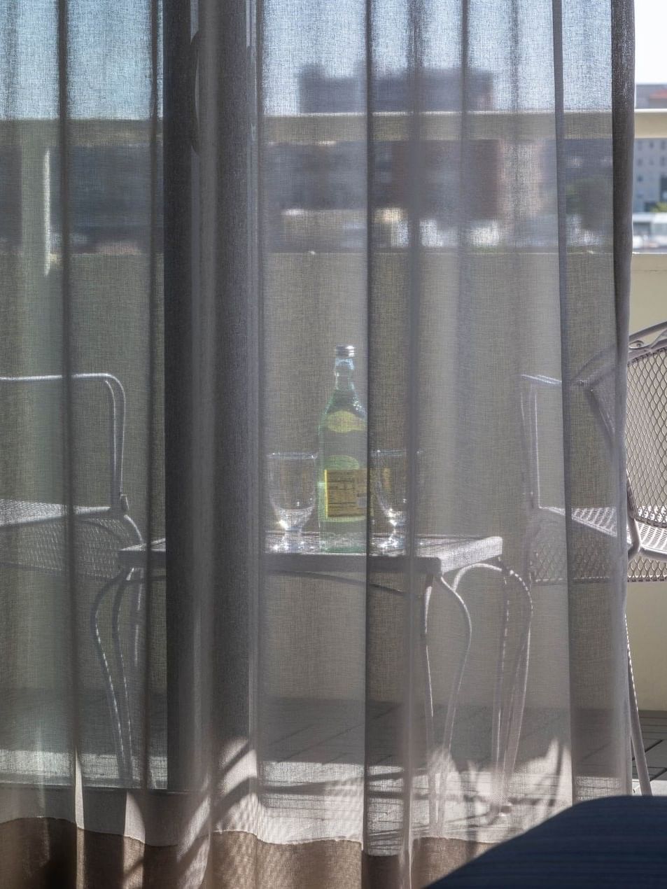 Balcony chairs by a table with water under a sunny sky through sheer curtains in Accessible Deluxe Rooms at Warwick Denver