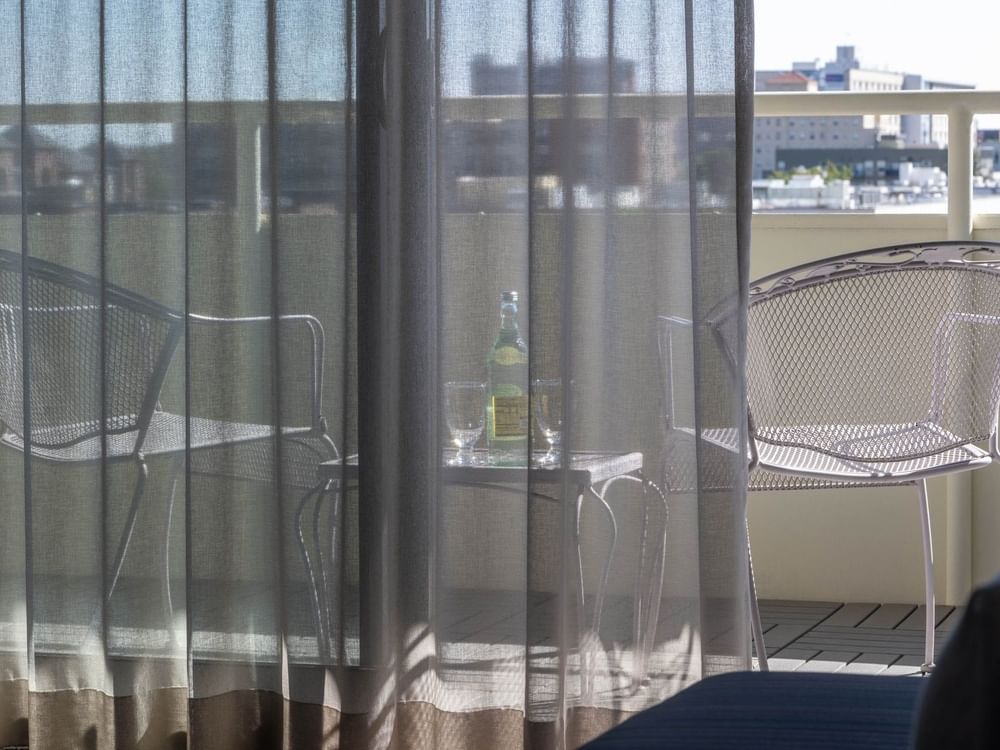 Balcony chairs by a table with water under a sunny sky through sheer curtains in Deluxe Rooms at Warwick Denver