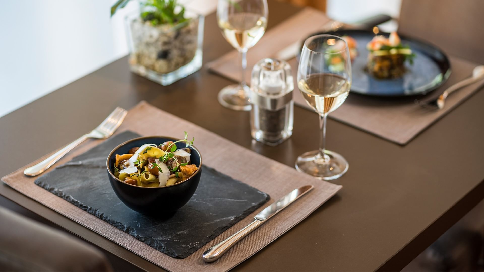 Two place settings with bowls of pasta, wine glasses, and utensils on a brown table.
