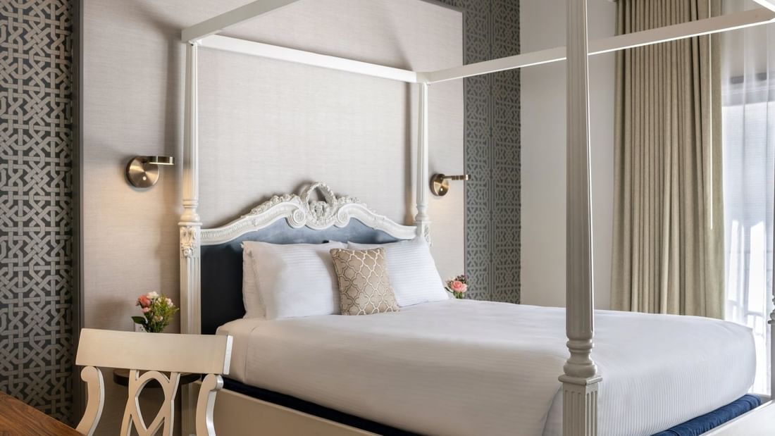 White four-poster bed by a carved desk chair under a grey patterned wall in Balcony Junior Suite at Warwick San Francisco