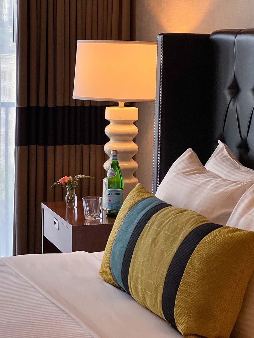 Close-up of a white bed with a colorful pillow by a lamp on a wooden nightstand in Queen Anne Suite at Warwick Seattle