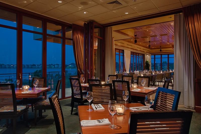 A dining area with wooden tables, chairs, wine glasses, and candles, surrounded by large windows.