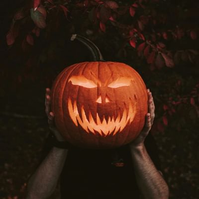 Mani che sostengono una zucca di Halloween intagliata con un volto spaventoso, illuminata da una luce calda.