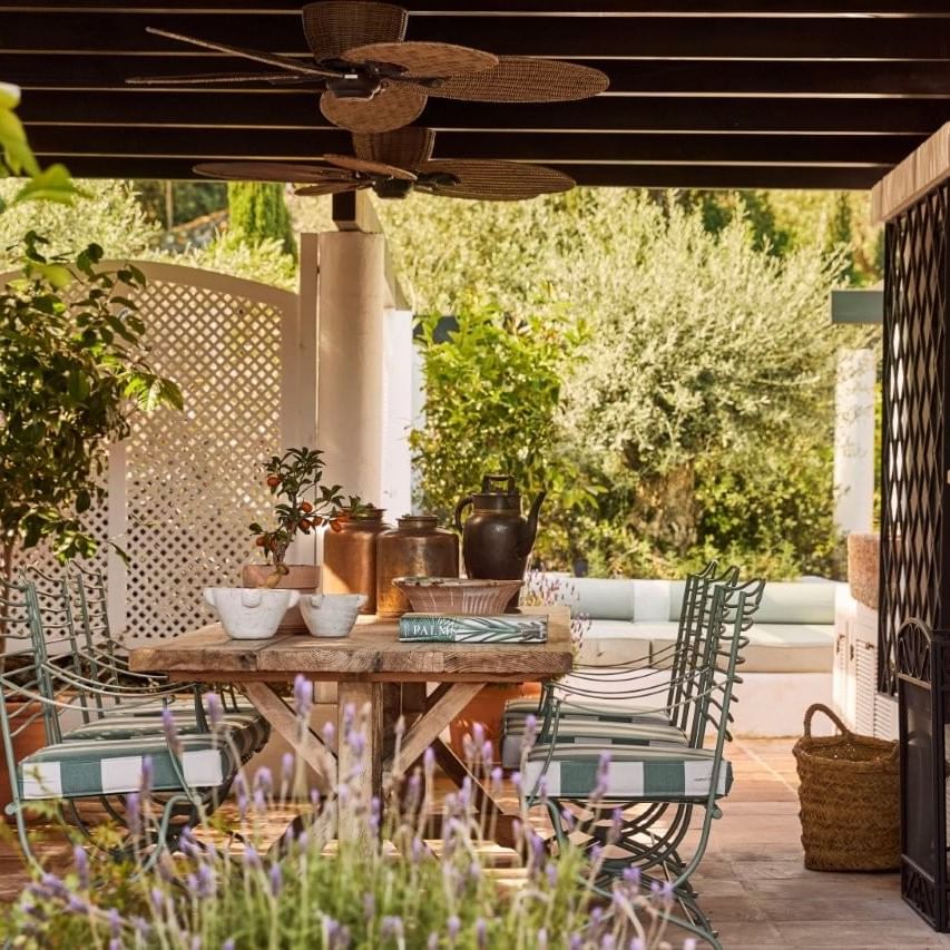 Patio with table, chairs, plants, fan, and distant greenery under a wooden ceiling.